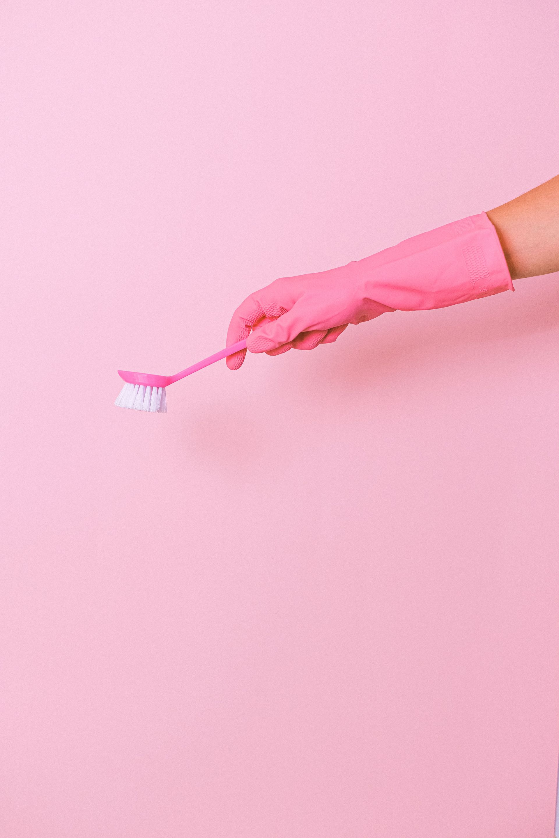 Unrecognizable cleaner wearing protective glove using plastic brush in hand to remove dirt during household against pink background in studio