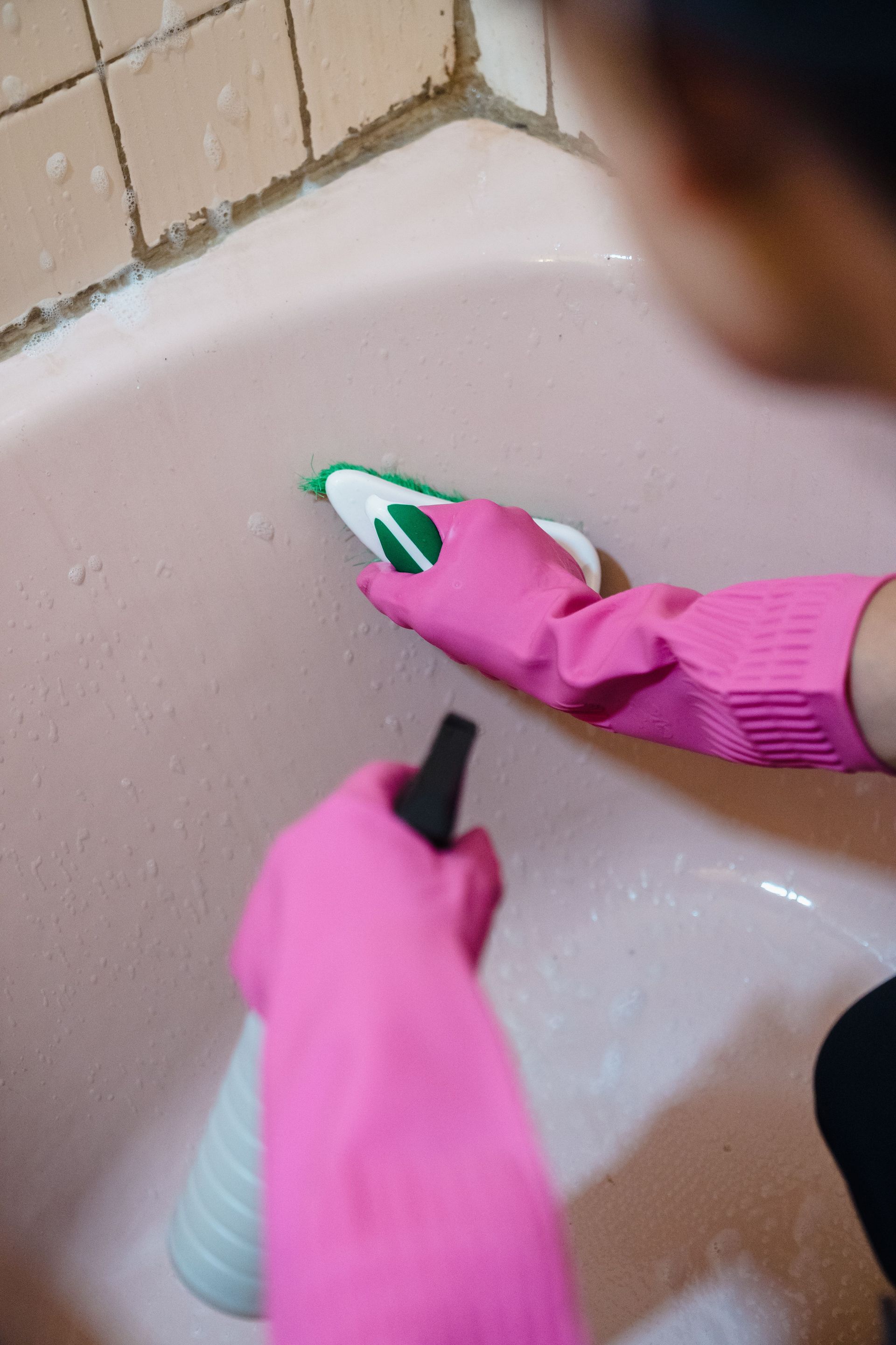 Person using pink gloves and scrubber to clean a bathtub with tiles.